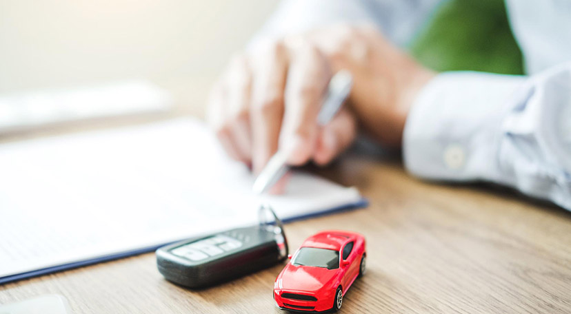 Car keys and a red toy car on a table, with man signing a car financing business loan in the background